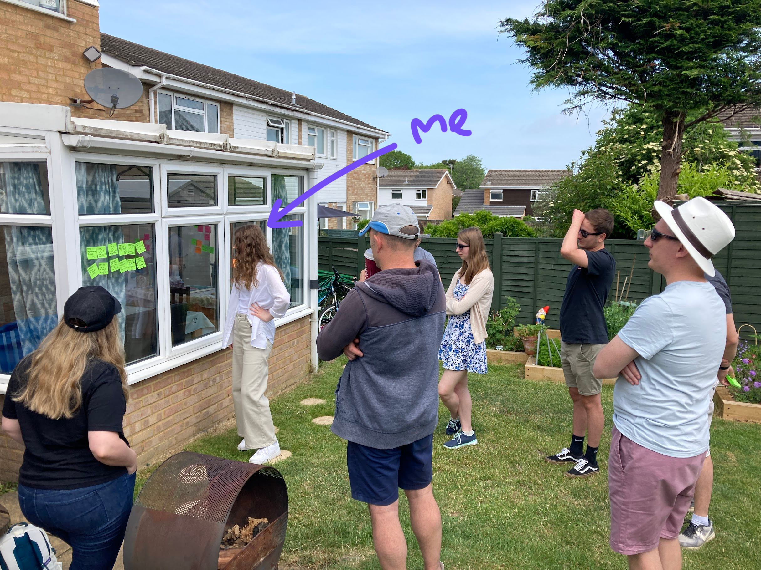 An image of a woman leading a workshop in a garden. There are several people listening to her, and sticky notes on a glass window in front of her.